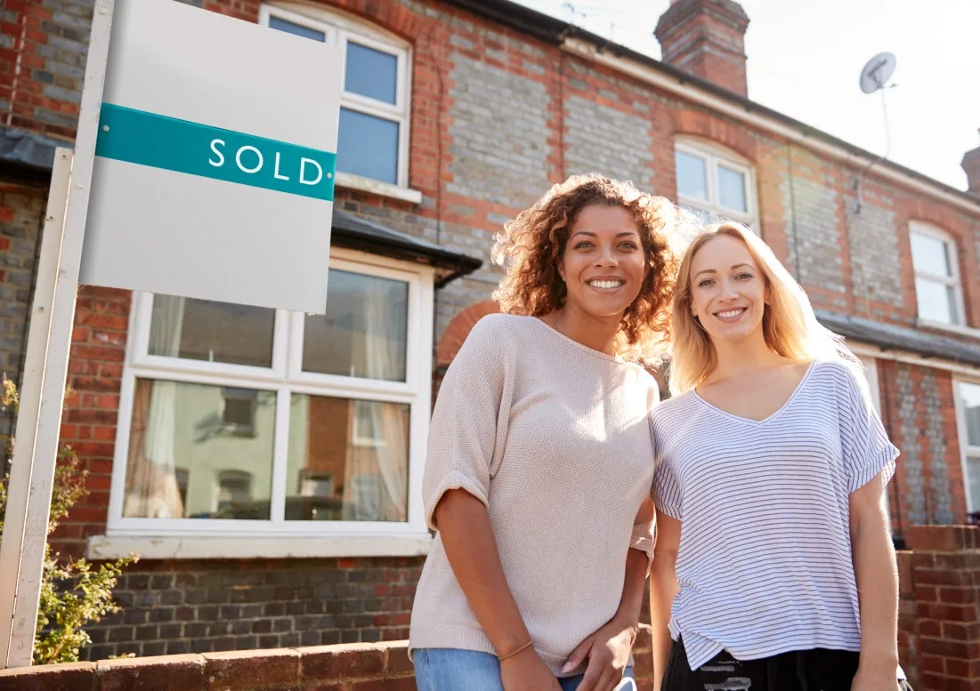 Two smiling women in front of a sold sign outside a house.